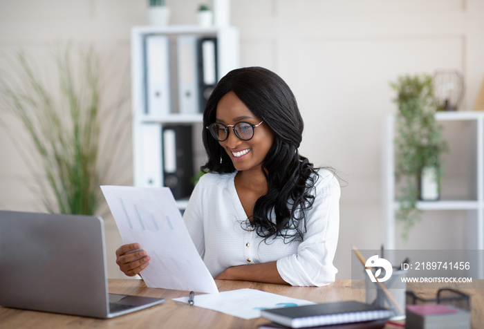 Attractive black businesswoman working with documents in front of laptop in modern office