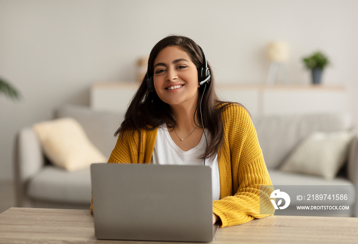 电子学习概念。快乐的阿拉伯女人戴着耳机，在家里用笔记本电脑在线学习，坐着