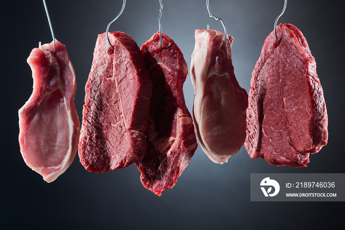 assorted raw meat steaks on metal hooks on dark black background