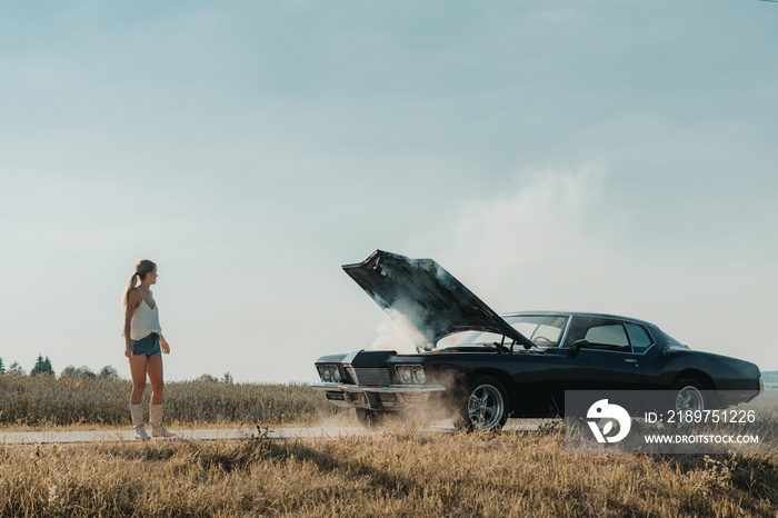 Young female standing near overheated car in the field, bright sunlight, steam under the hood