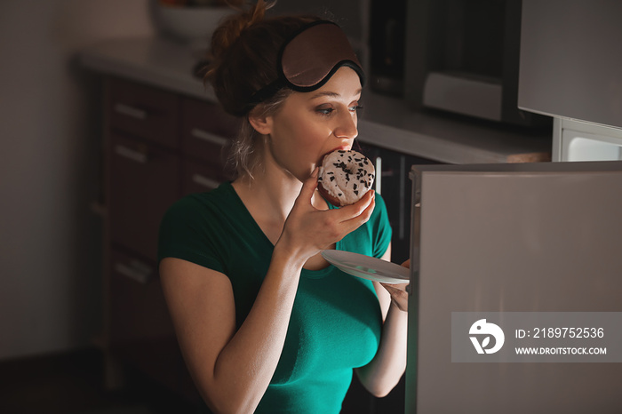 Beautiful young woman eating unhealthy food near refrigerator at night