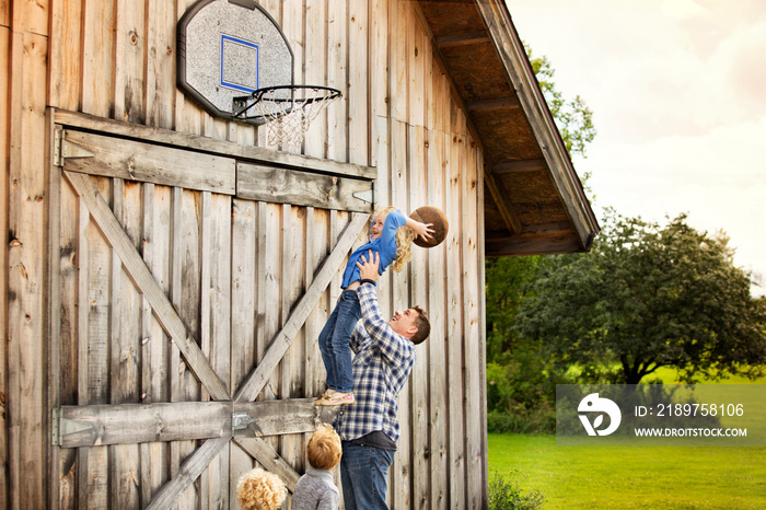 Father with children (2-3, 4-5, 6-7) throwing basketball ball into hoop fixed to barns wall