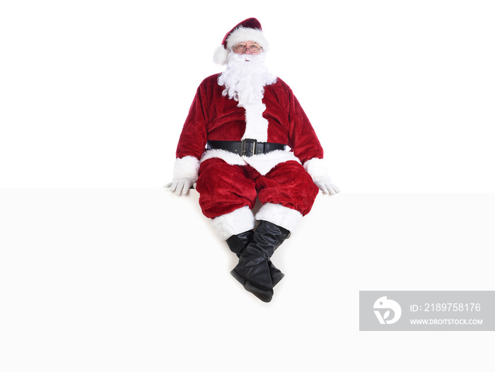 Senior man in traditional Santa Claus Suit sitting on a white wall.  Isolated on white with copy spa