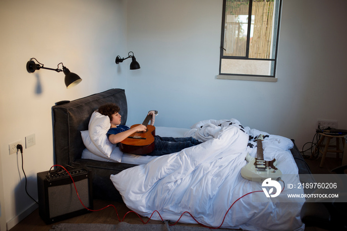 High angle view of boy playing guitar while relaxing on bed against wall at home