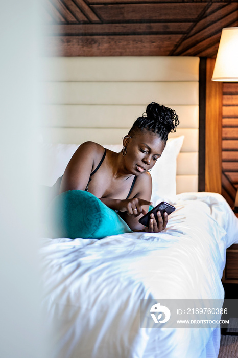 Black woman with braids laying on a blue green pillow in bed with cell phone