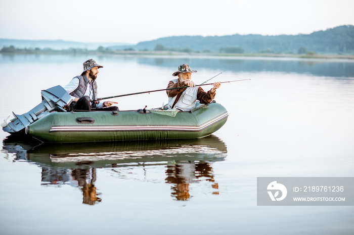 Grandfather with adult son fishing on the inflatable boat on the lake early in the morning