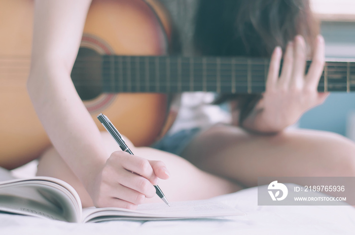 Young beautiful woman sitting on her bed in the bedroom holding guitar composing a song  and writing