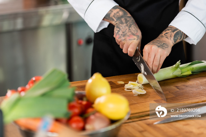 纹身厨师在厨房模糊的蔬菜附近割韭菜的裁剪图