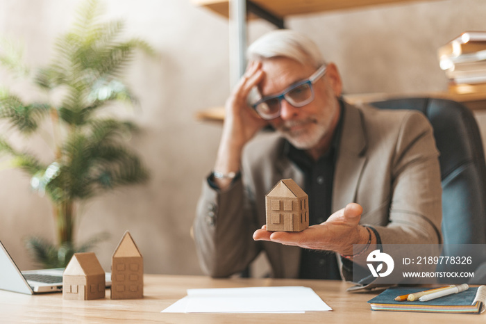 Real estate crisis. Mature man in the office at the table holding a model of the house. Financial ad