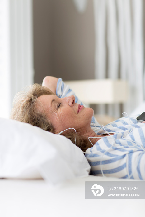 Serene senior woman listening to music with headphones on bed