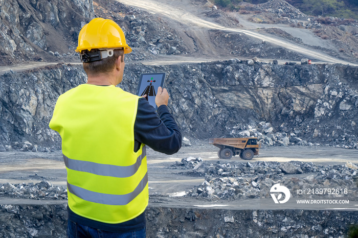 An engineer holds a tablet in his hand and remotely controls a heavy-duty dump truck in the backgrou