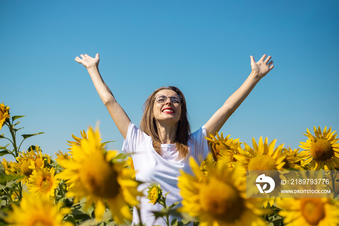 Young woman in a white t-shirt and glasses with raised up hands on a sunflower field on a summer sun