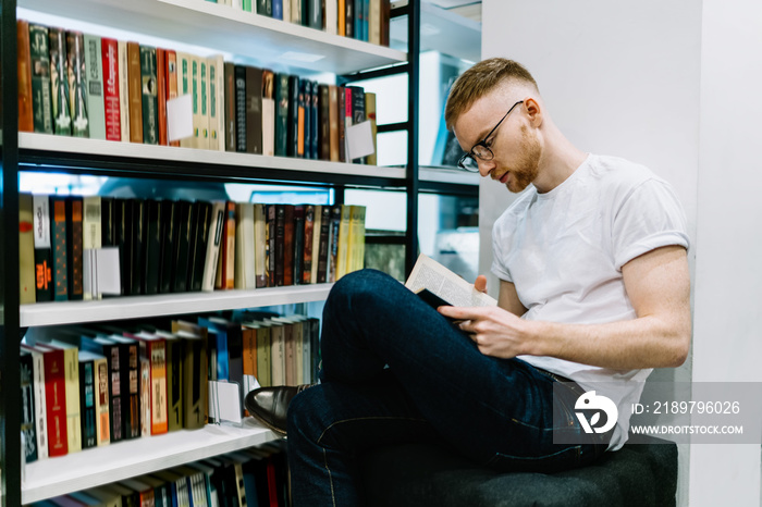 Pensive student sitting cross legged and reading book in library