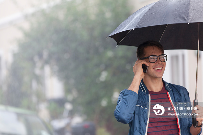 Smiling young man with umbrella talking on smart phone in rain
