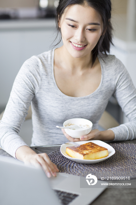 年轻女子一边吃早餐一边使用笔记本电脑