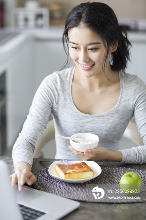 年轻女子一边吃早餐一边使用笔记本电脑