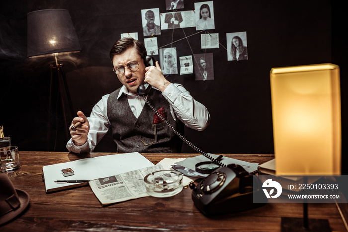 Irritated detective in glasses holding cigar and talking on telephone in dark office