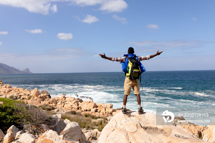 Fit afrcan american man wearing backpack hiking spreading arms on the coast