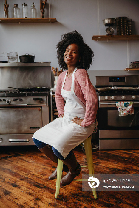 Portrait of smiling female chef with curly hair sitting on stool against appliances in cooking class