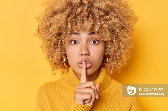 Headshot of surprised curly haired young woman presses index finger over lips makes shush gesture lo