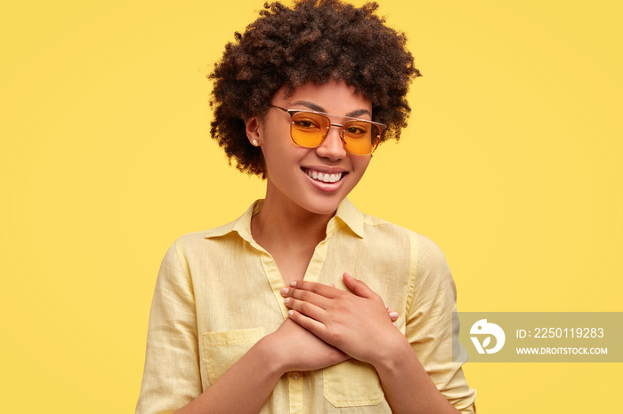 Positive glad lovely young woman with Afro hairstyle, keeps hands on heart, expresses her sincere em