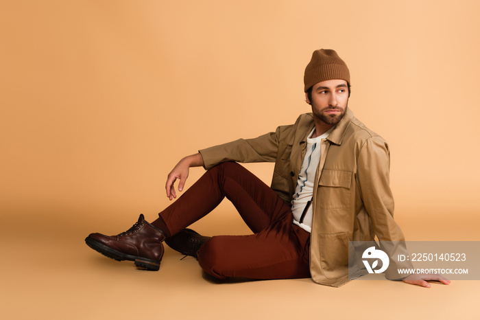 trendy man in stylish autumn outfit looking away while sitting on beige