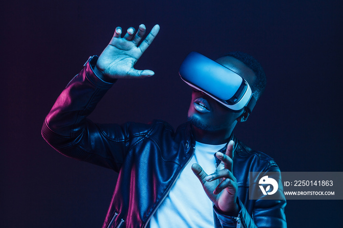 Young african man wearing virtual reality goggles with hands up, isolated on black background