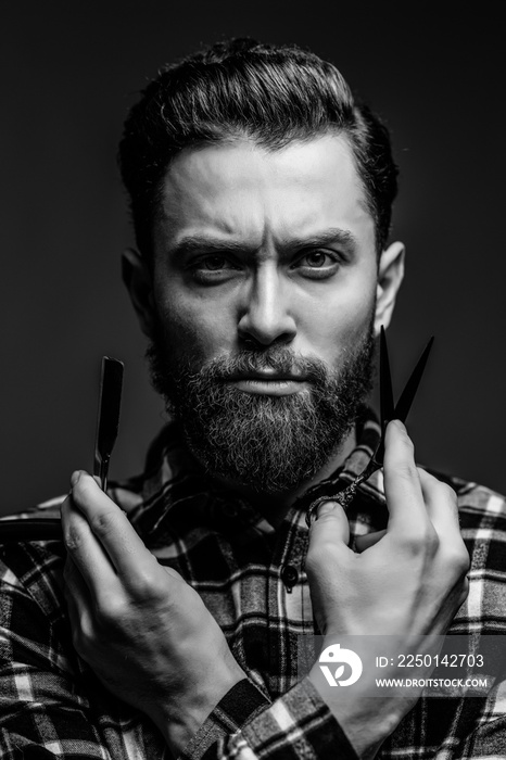 Portrait of bearded youngman with barber scissors and straight razor standing isolated on gray backg