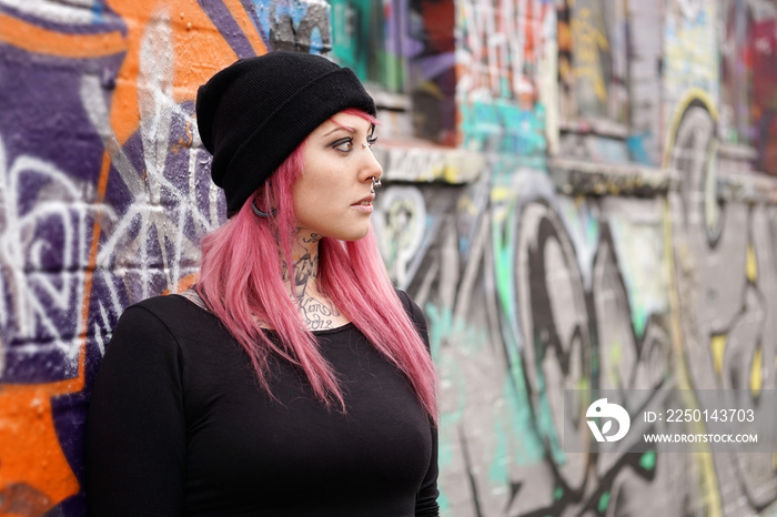 年轻的另类女性，粉色头发穿孔，纹身靠在涂鸦墙上