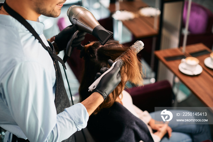 Close-up of hairdresser uses round brush while styling womans hair at hair salon.