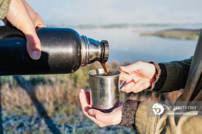 Pouring hot coffee out of thermos flask at beautiful riverbank. Two hikers enjoy a morning drink on 