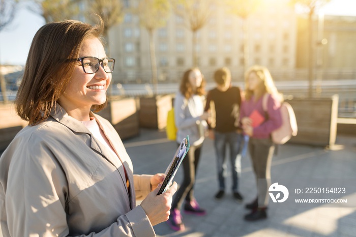 Portrait of mature smiling female teacher in glasses with clipboard, outdor with a group of teenager