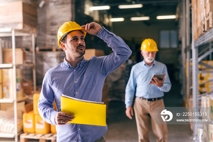 Businessman holding helmet and folder with documents while looking up and standing in warehouse. In 