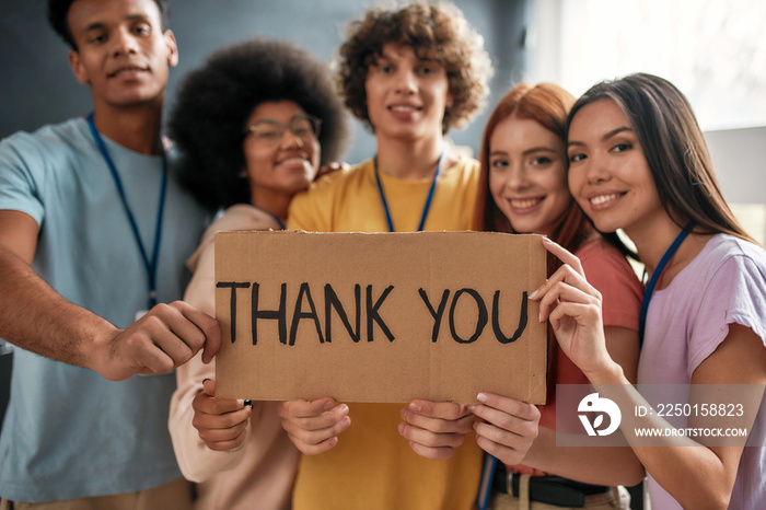 Group of diverse young volunteers smiling at camera, holding card with Thank you lettering while sta