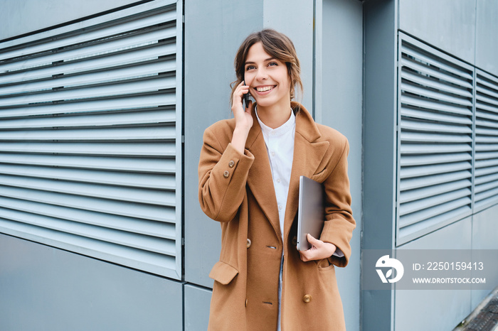 Young cheerful businesswoman in coat with laptop happily looking away while talking on cellphone out