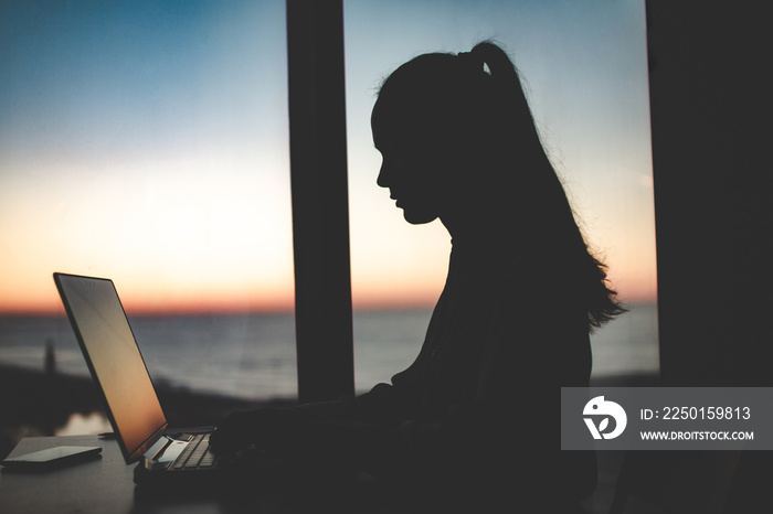 Silhouette of a business woman working late at a laptop against the window at sunset. Night and even