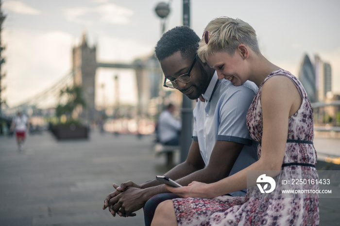 Young couple sitting on wall, looking at smartphone, Tower Bridge in background, London, England, UK