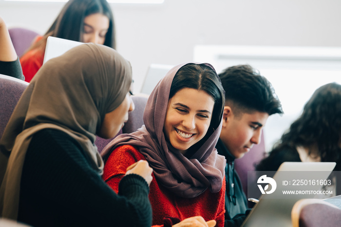 Smiling female classmates discussing over laptop during university lectures