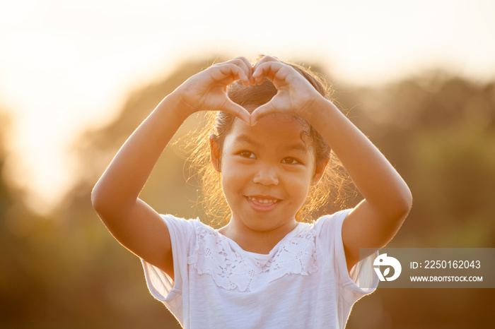 Cute asian child girl making heart shape with hands in the field with sunlight