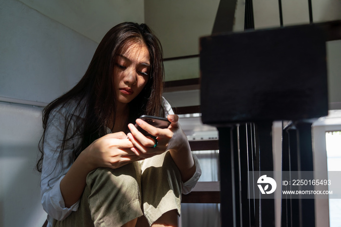 选择性关注孤独年轻漂亮的亚洲女人坐在家里的楼梯上使用智能手机。