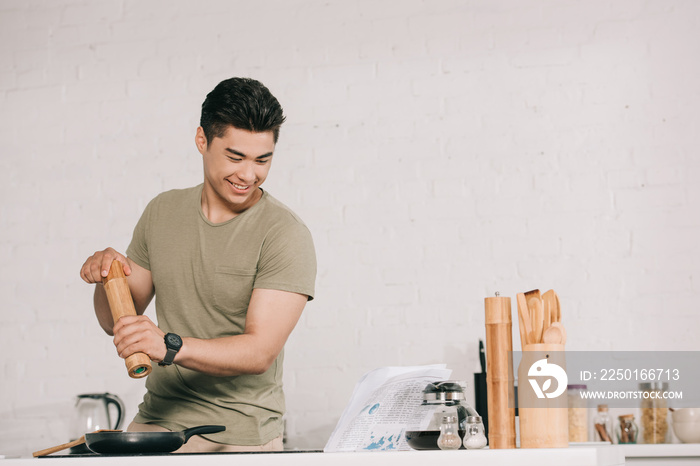 cheerful asian man cooking breakfast while reading newspaper in kitchen