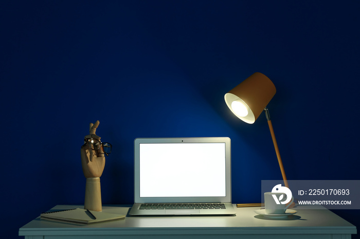 Modern workplace with open laptop and glowing lamp near blue wall in dark room