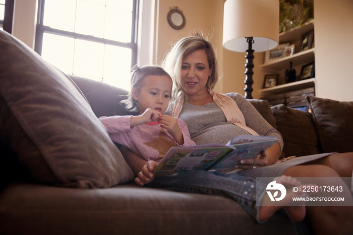 Low angle view of pregnant mother with daughter reading book while sitting on sofa at home