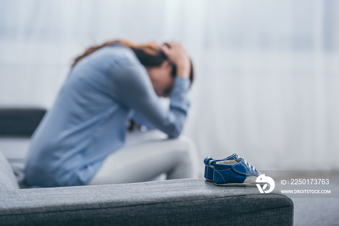 selective focus of blue baby shoes on couch with sad woman sitting and lowering head at home, grievi