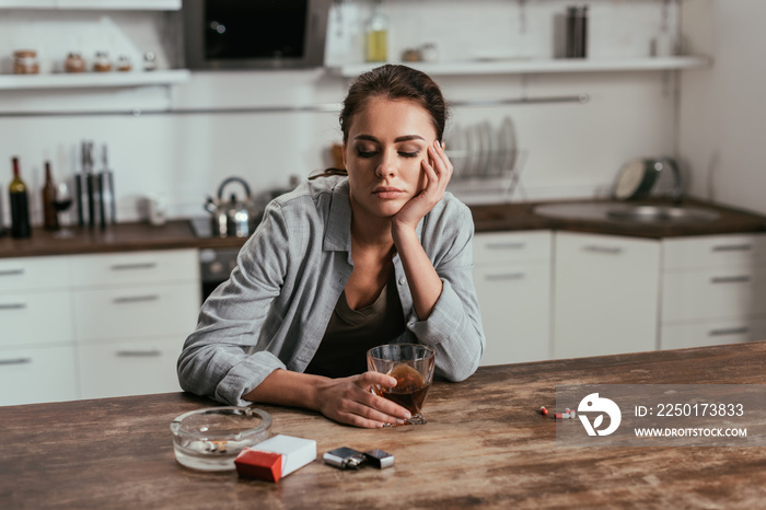 忧心忡忡的女人拿着威士忌酒杯，旁边放着桌子上的香烟和药片