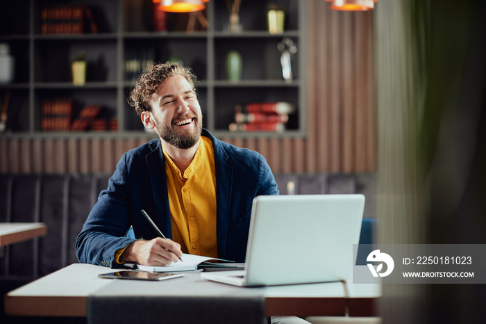 Young smiling bearded Caucasian blogger dressed smart casual writing notes in agenda and looking at 