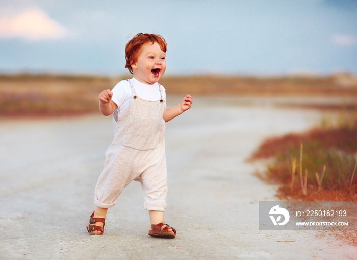 adorable redhead toddler baby boy in jumpsuit running through the summer road and field