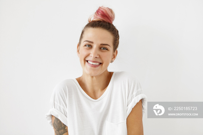 Glad blue-eyed female with pink hair knot, wearing loose white shirt, having tattooes on arms, smili