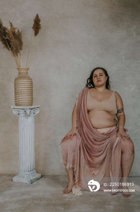 plus size metis woman wearing pink fabric and nude bra and underwear sitting next to roman column