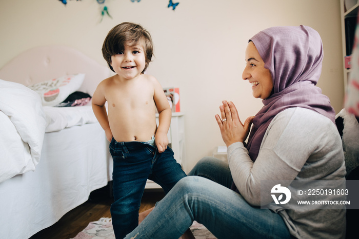 Smiling mother cheering little boy wearing jeans in bedroom at home